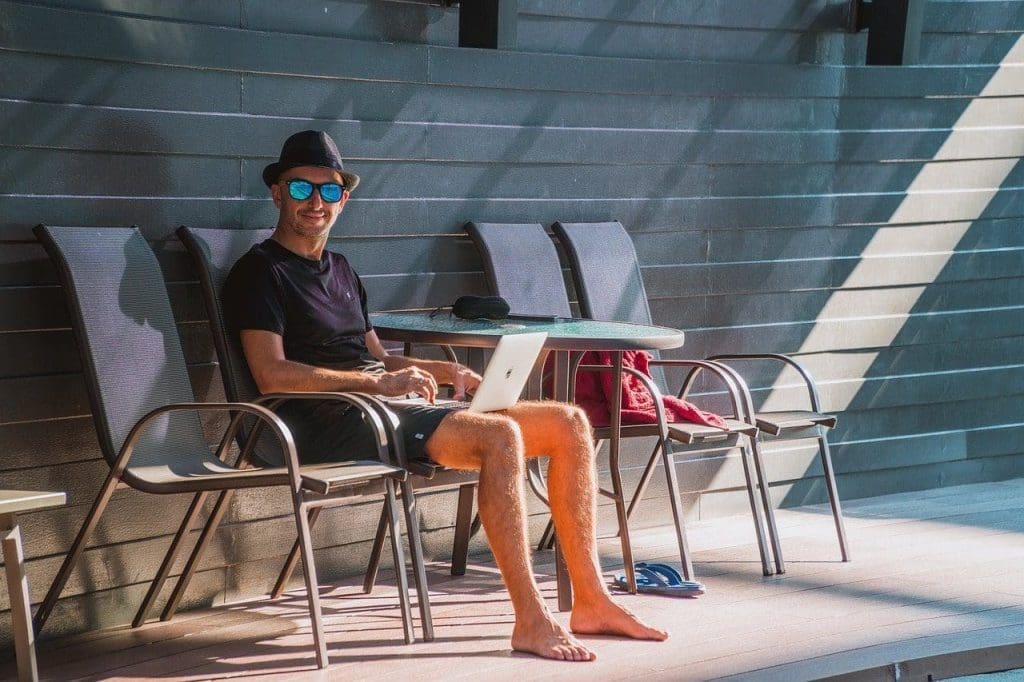 man sitting outside at laptop wearing hat and sunglasses shorts