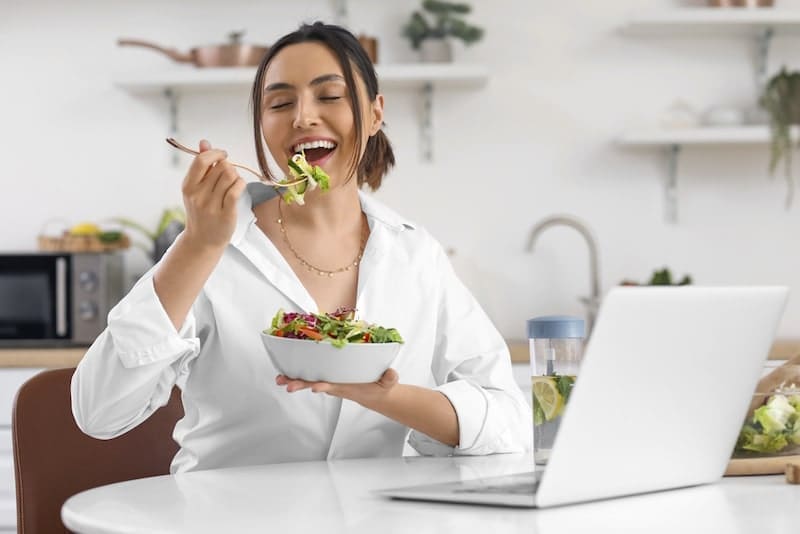 Young woman eating vegetable salad at table in kitchen